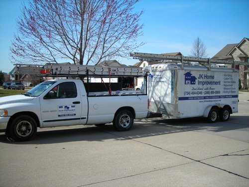 A white pickup truck with ladders on top is towing a trailer labeled JK Home Improvement in a residential neighborhood. The trailer displays contact information and services offered by the company.