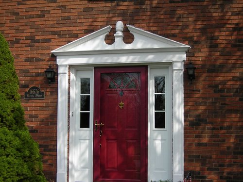 A red front door with a brass knocker and handle is set in a white-trimmed entrance with two narrow side windows, on a brick house. A green shrub is on the left, and a house number plaque reads 16576 nearby.