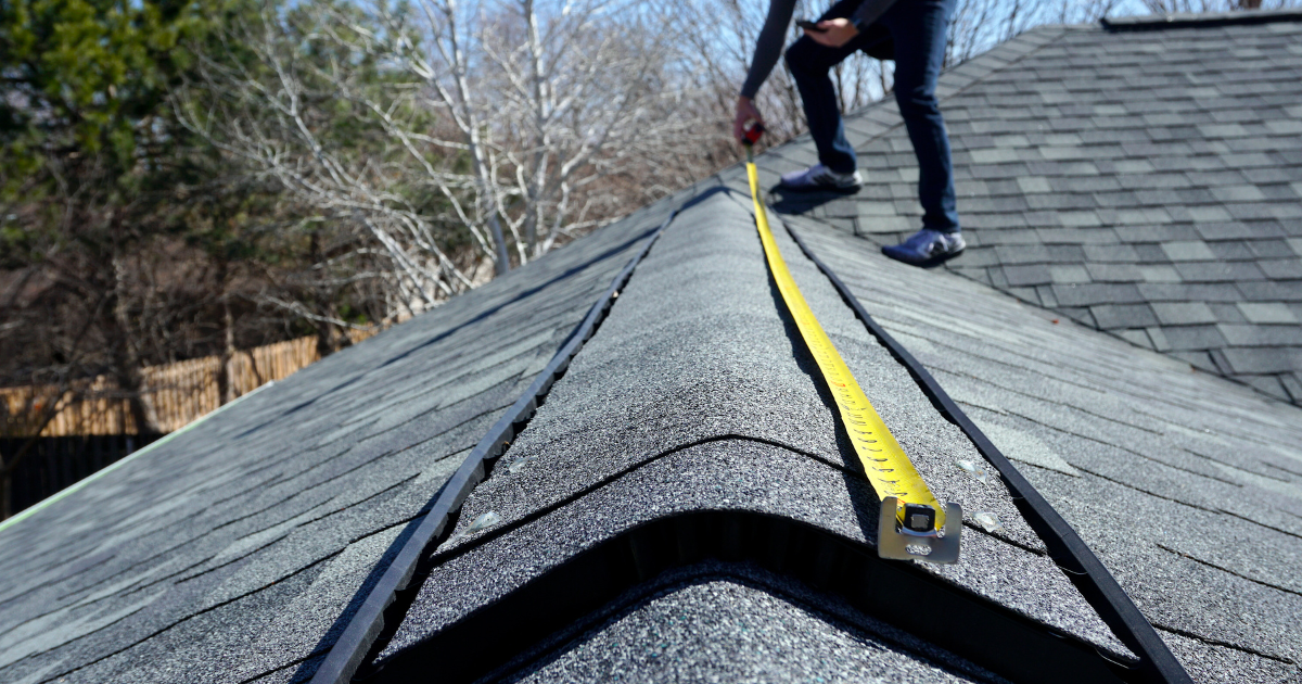 Roofing (2) A person stands on a shingled roof, using a yellow measuring tape to measure along the roof ridge on a clear day. Bare trees and a wooden fence are visible in the background.