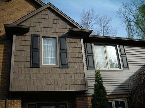 A close-up of a house exterior shows two second-story windows with black shutters, brown wood shingles, and beige siding. A small evergreen tree is visible near the base of the house.