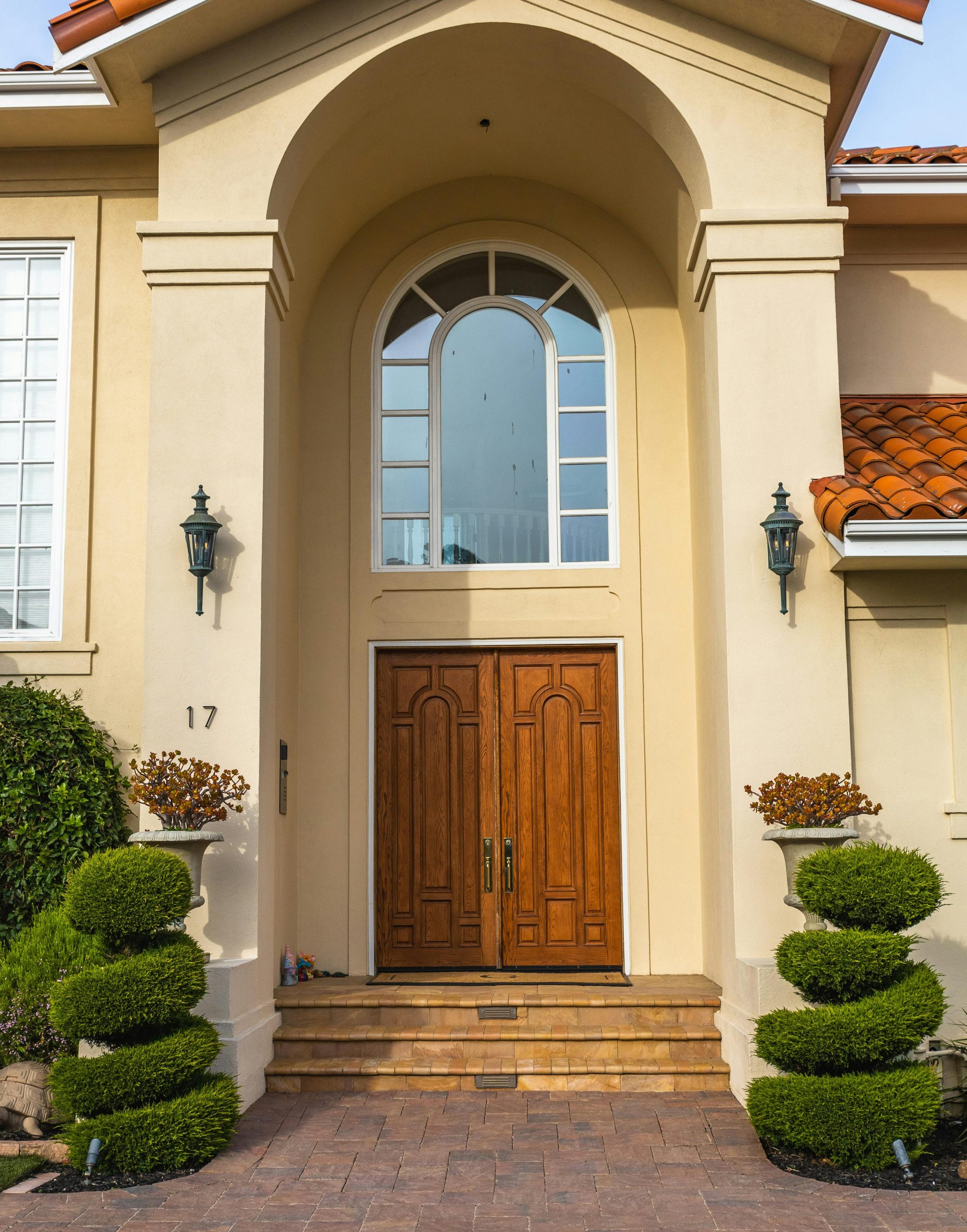 Front entrance of a beige house with an arched window above double wooden doors, flanked by two lantern-style lights and symmetrical trimmed topiary bushes in planters on either side of the steps.