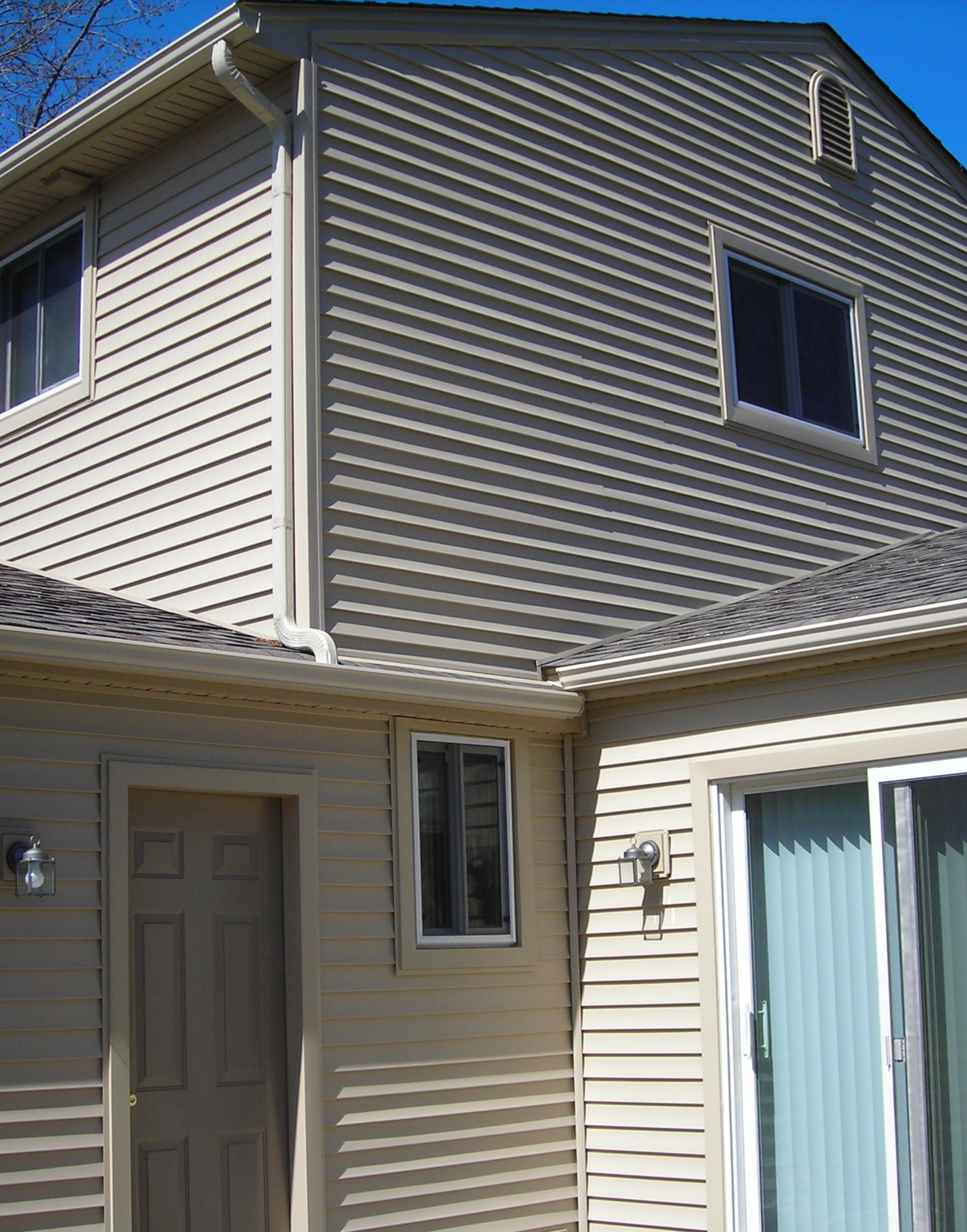 Beige vinyl-sided house exterior with several windows, a tan door, an outdoor light fixture, a sliding glass door, and visible gutters under a clear blue sky.