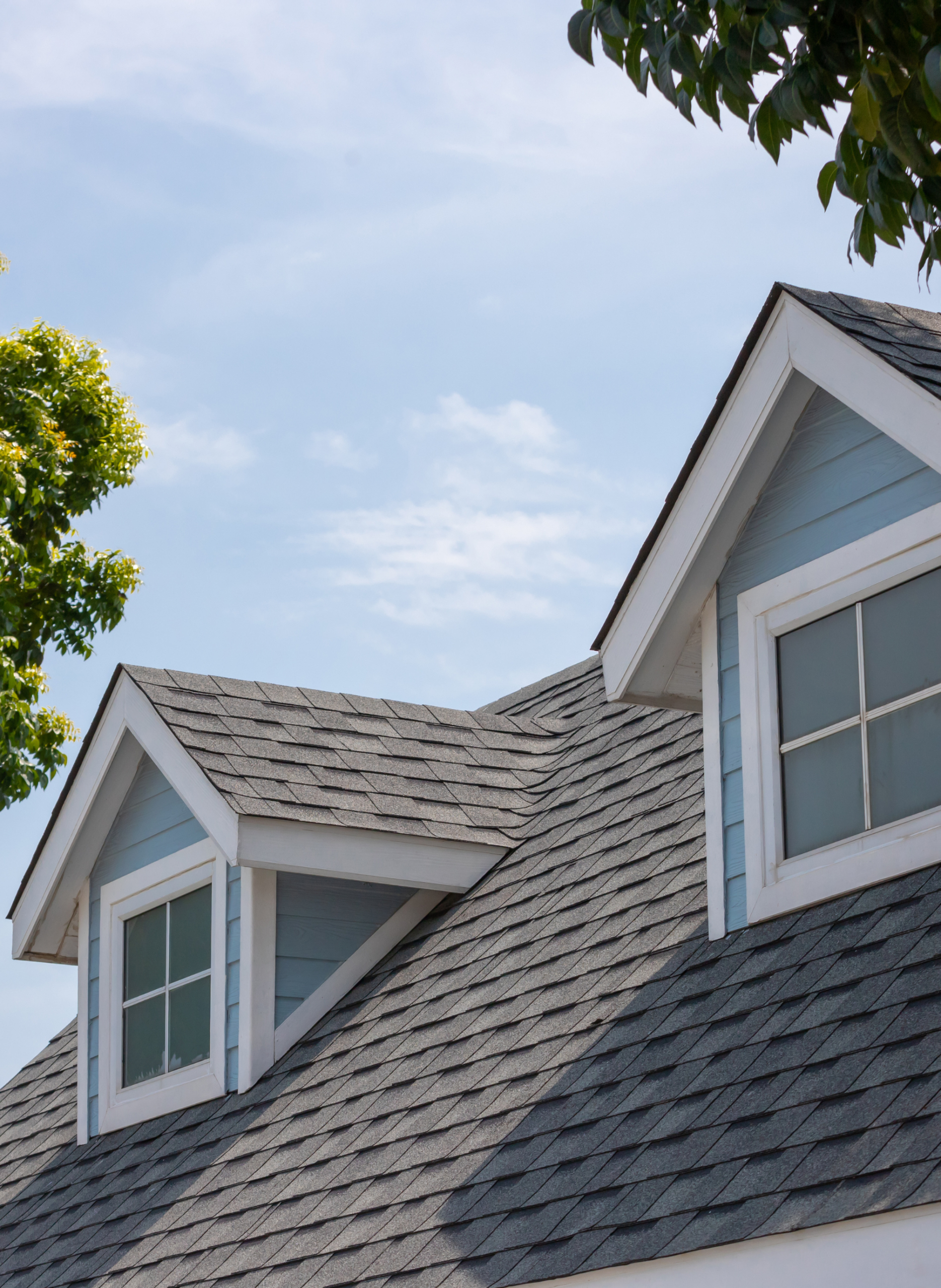 roofing A close-up view of a house roof with gray shingles, featuring two dormer windows with white trim against light blue siding, surrounded by green tree branches under a blue sky.